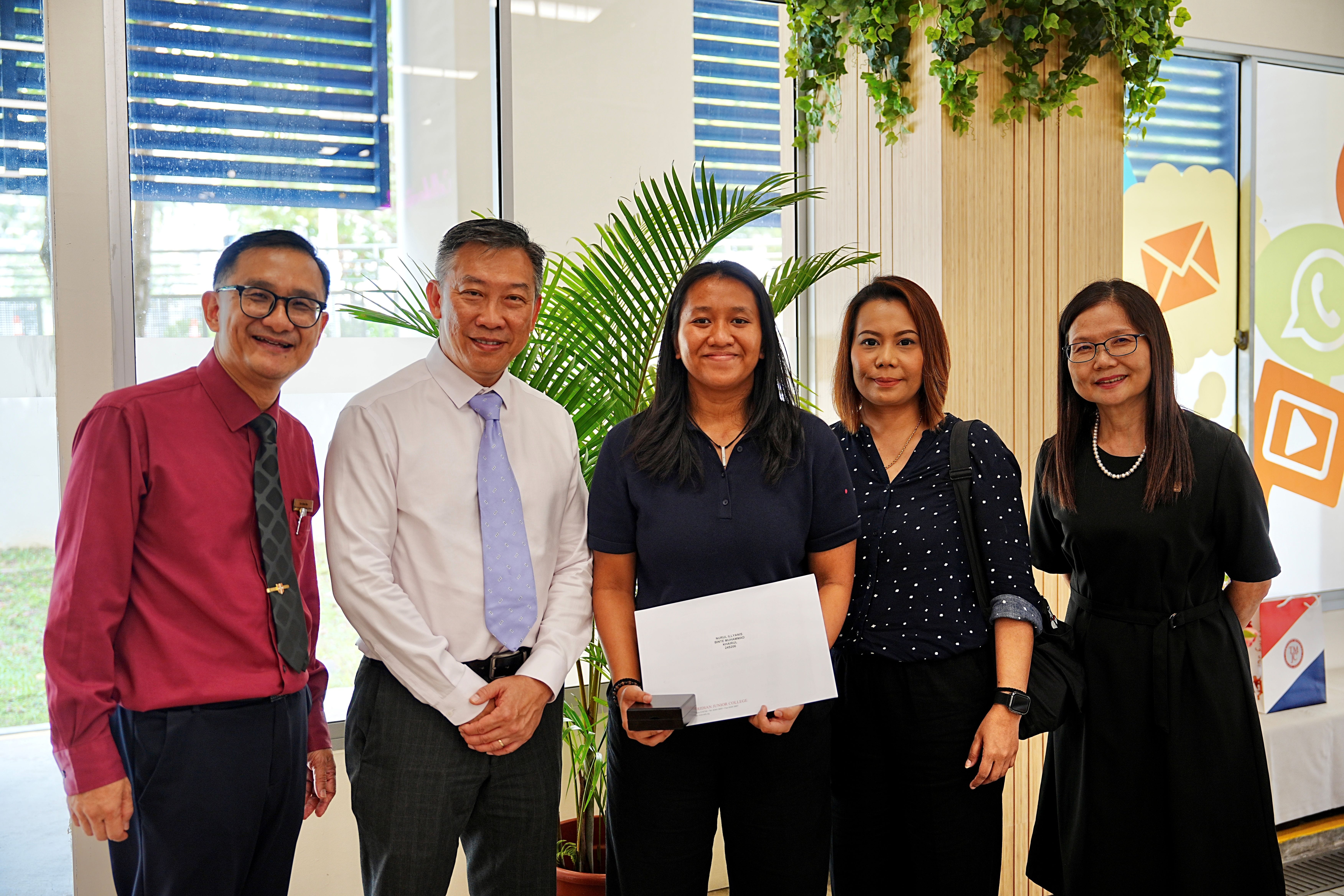 Five people stand together indoors for a photo. The person in the centre, a woman in a dark polo shirt, holds an envelope and a small box, suggesting she is receiving an award. Two men stand to her left and two women to her right.